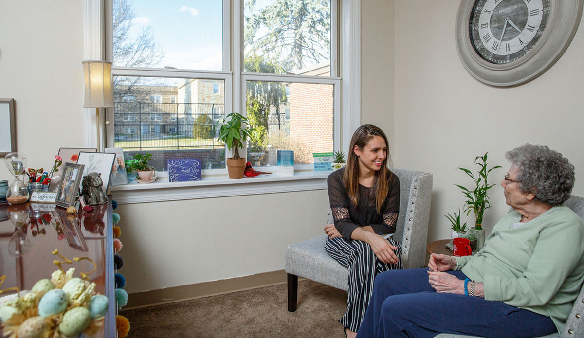 A woman sitting on a chair, engaged in conversation with another woman nearby.