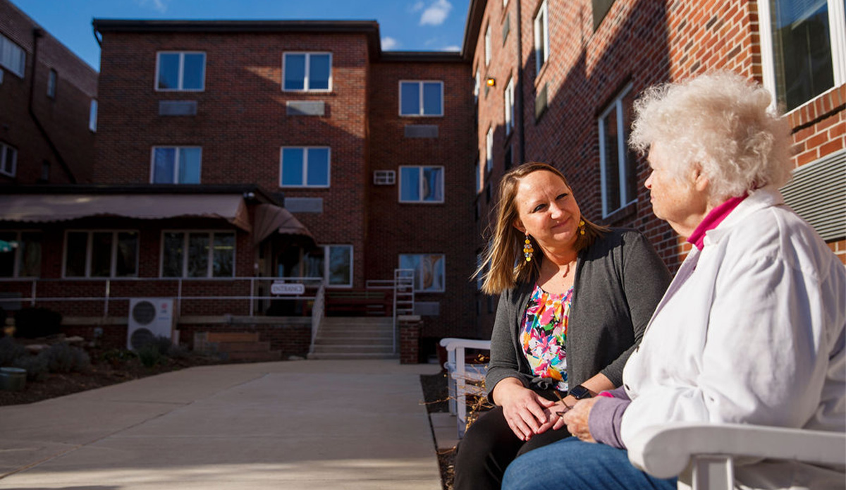 A woman converses with an older woman seated in a chair, sharing a warm moment together.